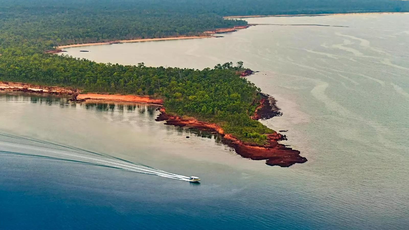 Aerial view of a boat on calm water near a forested coastal bay at sunset.
