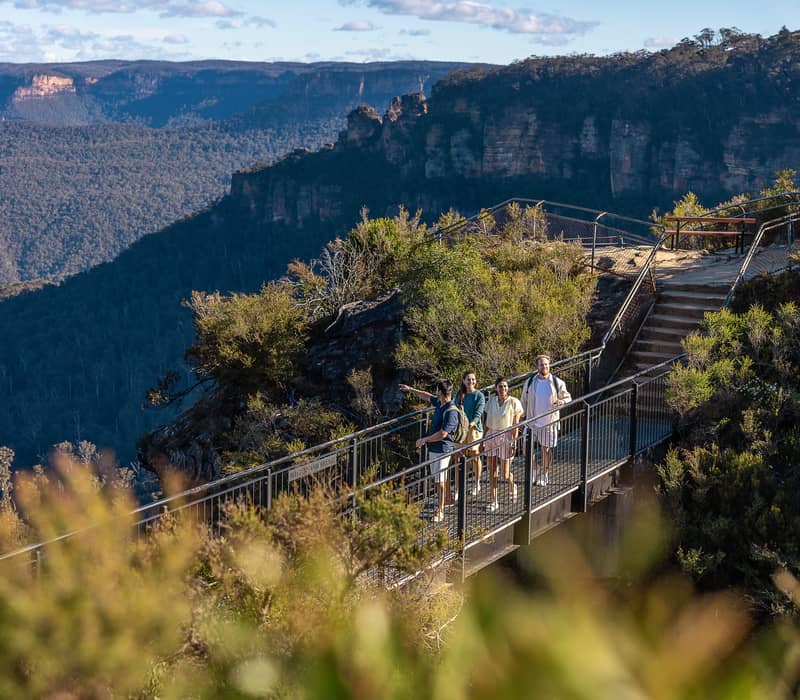 A group of four people walking across a metal bridge on a hiking trail overlooking the Three Sisters in the Blue Mountains.