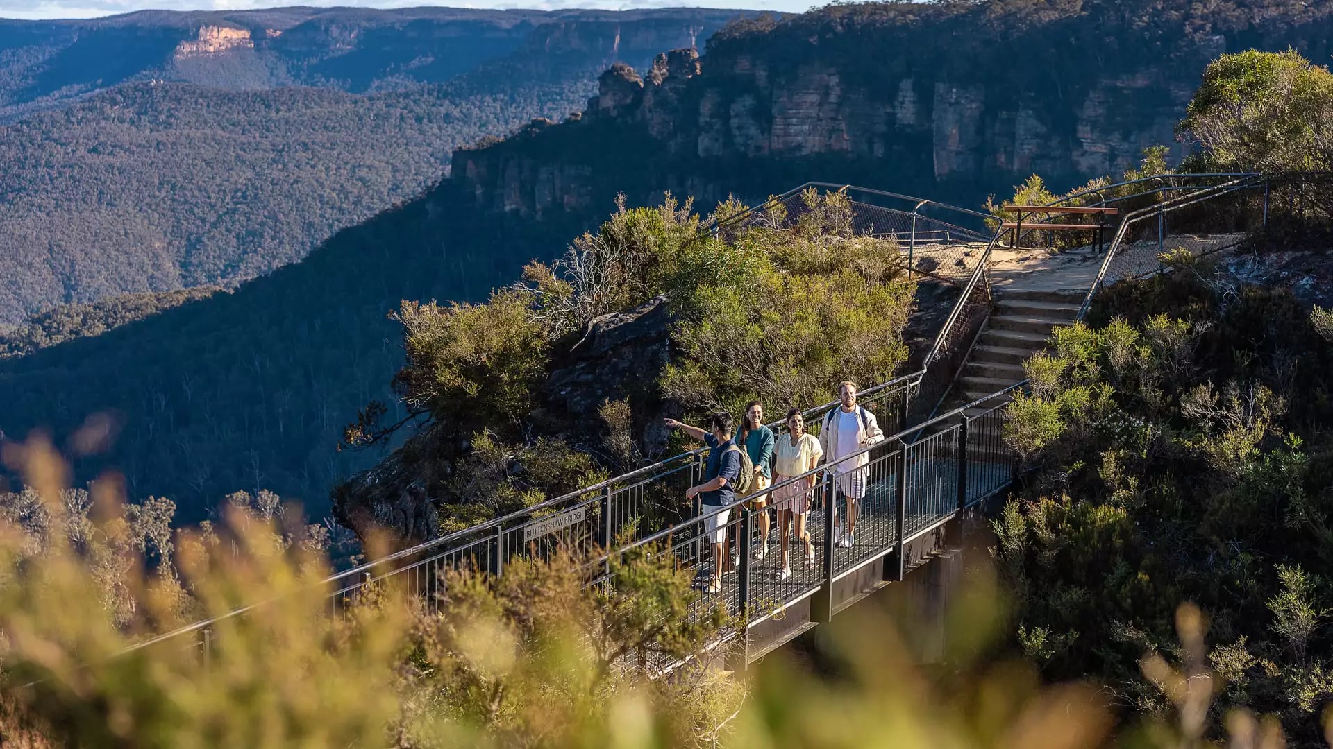 A group of four people walking across a metal bridge on a hiking trail overlooking the Three Sisters in the Blue Mountains.