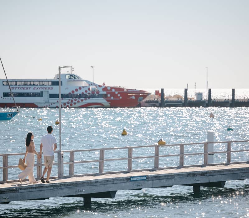 A man and woman walking on a pier over sparkling water with a red and white ferry in the background.