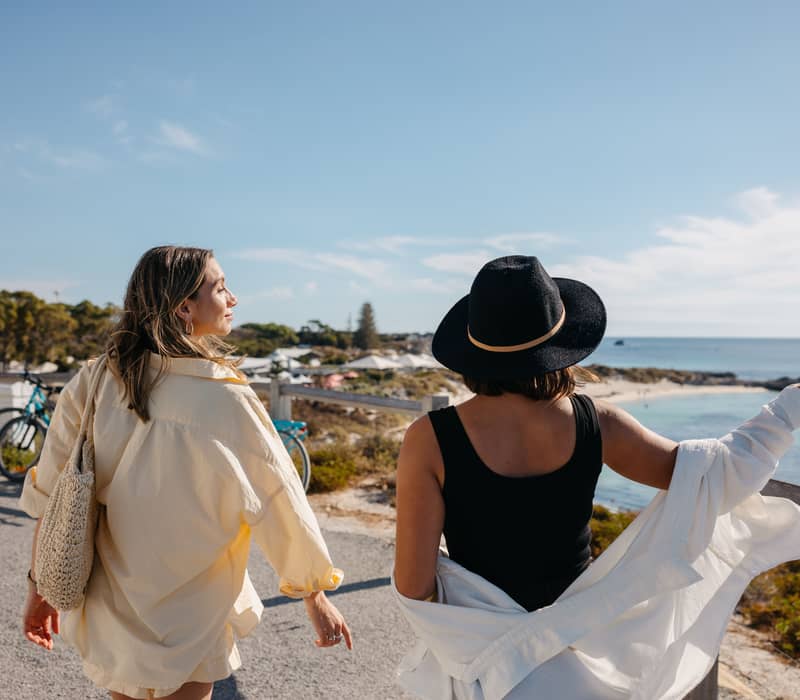 Two women standing on a path looking out over the blue ocean and coastline at Pinky's Beach, Rottnest Island.