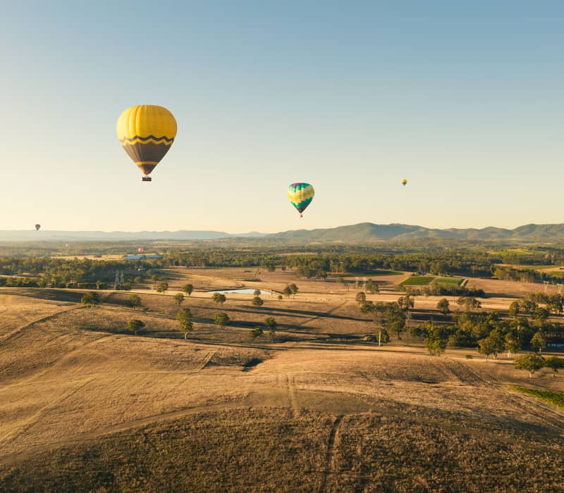 Multiple hot air balloons flying over the rural landscape and vineyards of the Hunter Valley, NSW.