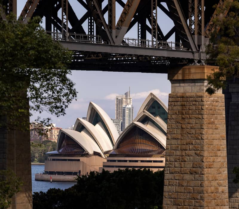 A view of the Sydney Opera House white sails seen through the pylons and beams of the Sydney Harbour Bridge.