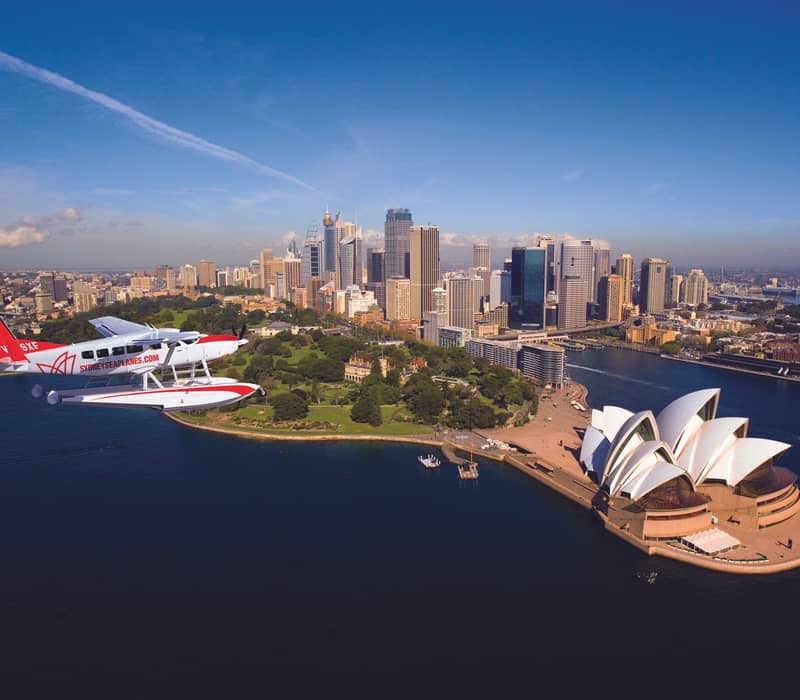 A red and white seaplane in flight over the Sydney Opera House and city skyscrapers.