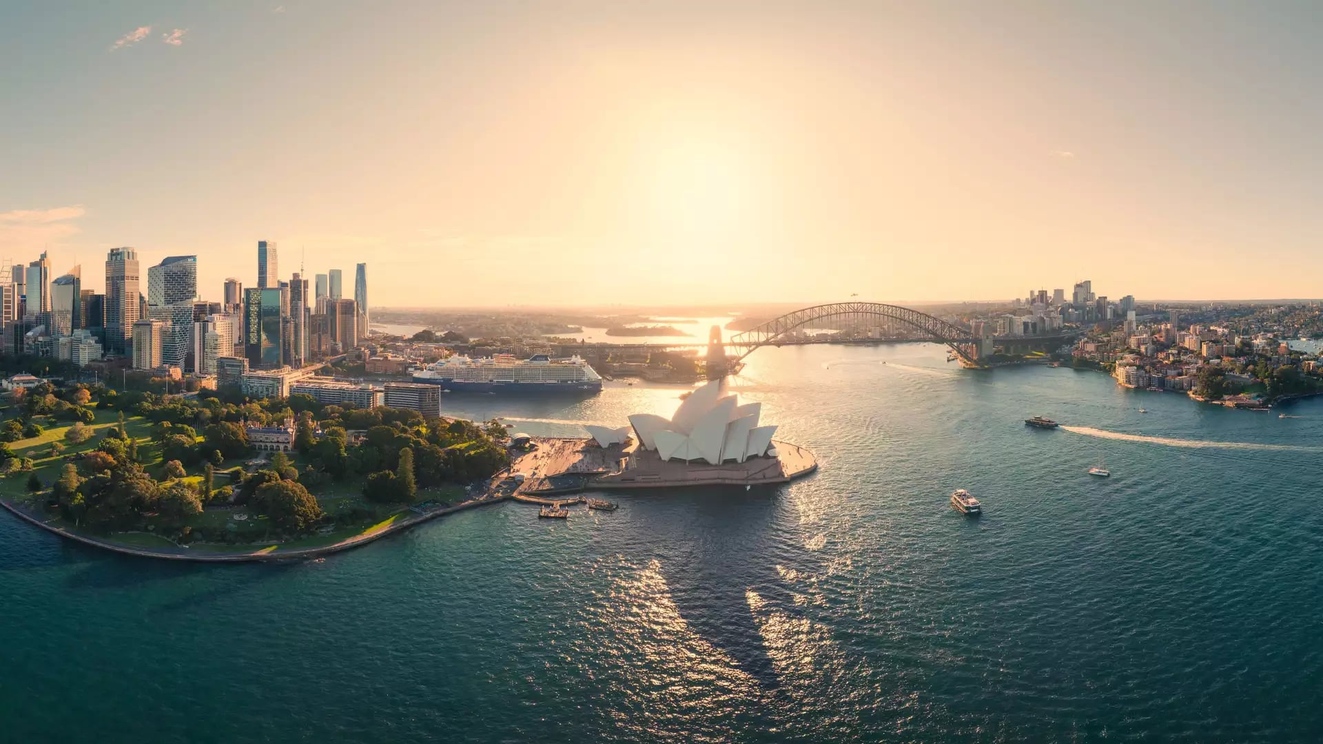 Panoramic aerial view of Sydney Harbour, the city skyline, and the Opera House under a golden sun.