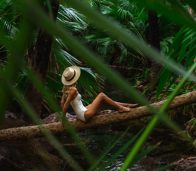 A woman in a white swimsuit and straw hat sits on a log over a shallow, clear stream deep within a dense, green tropical palm forest.