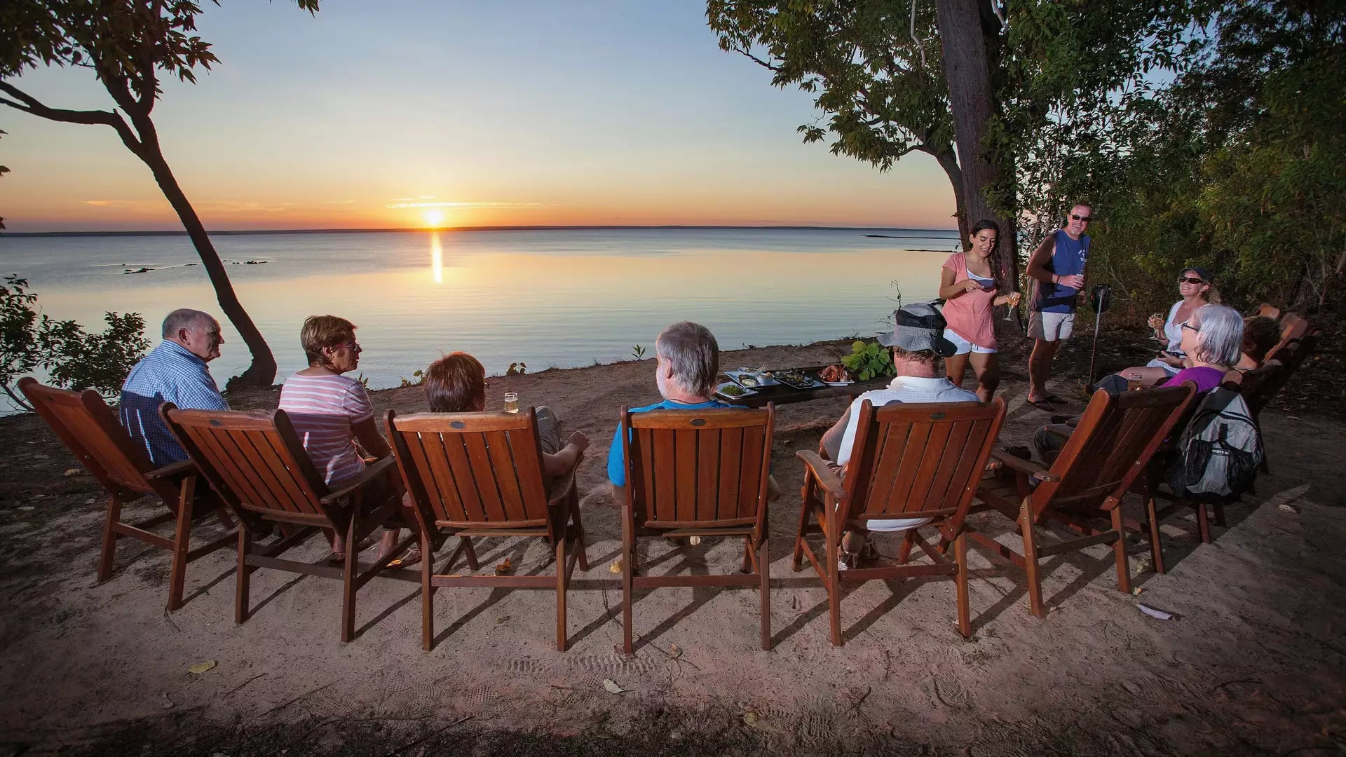 Travelers sitting in wooden chairs watching a sunset over water in the Top End.