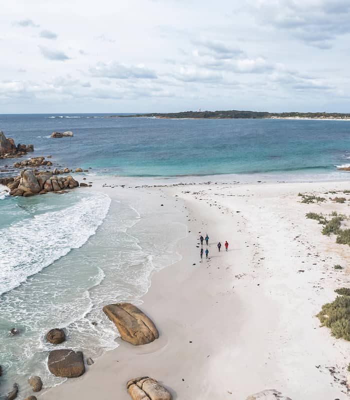 Aerial view of a small group of people walking along a white sand beach next to turquoise ocean and orange boulders.