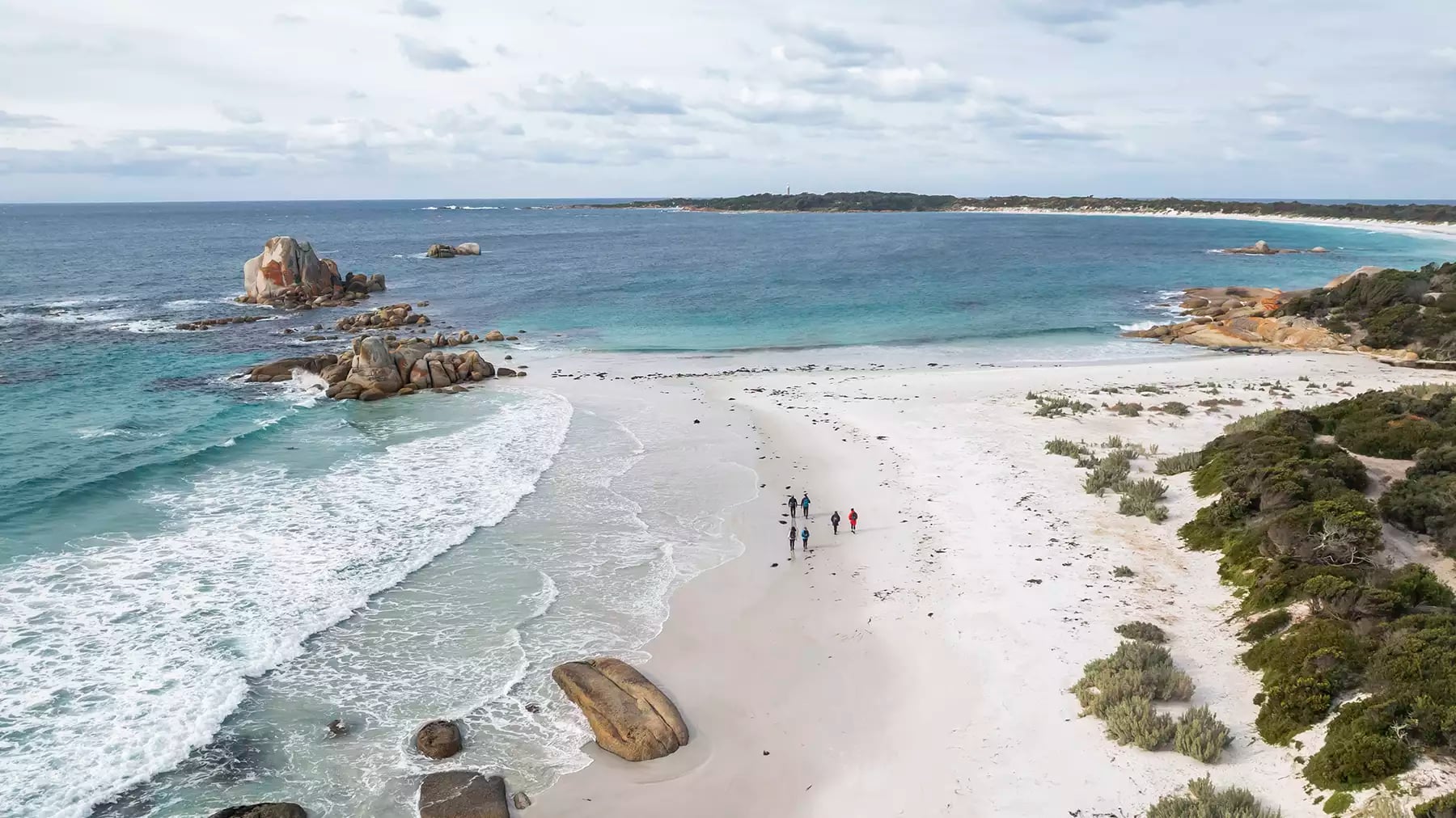 Aerial view of a small group of people walking along a white sand beach next to turquoise ocean and orange boulders.