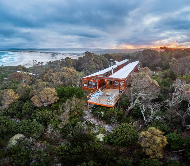 Aerial view of a long, modern timber lodge with a large deck overlooking the ocean at sunset.