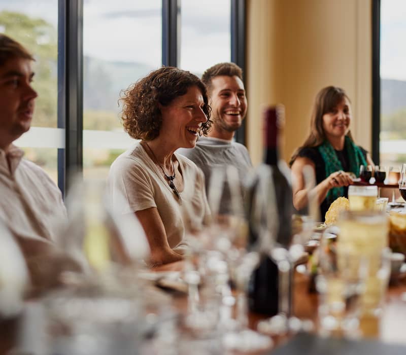 A group of adults laughing and talking at a rustic timber bar during a wine tasting at Fallen Giants.