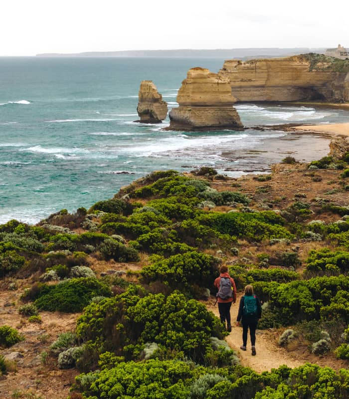 Two hikers walking on a dirt path through coastal vegetation toward sea stacks in the ocean.