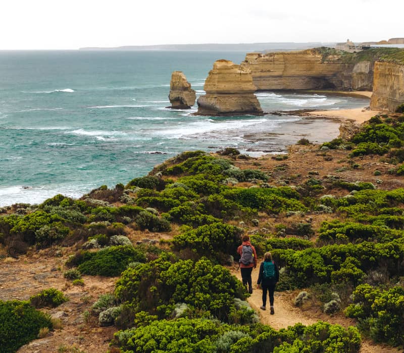Two hikers walking on a dirt path through coastal vegetation toward sea stacks in the ocean.