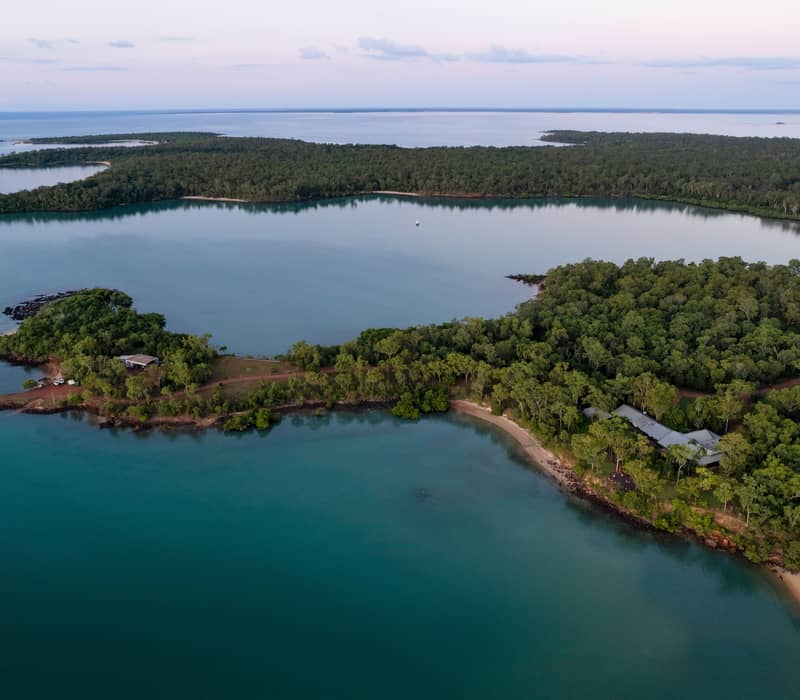 A high-angle aerial view showcases the secluded luxury of Seven Spirit Bay Wilderness Lodge, nestled within the pristine tropical forest of the Cobourg Peninsula. The image captures the lodge's unique position inside Garig Gunak Barlu National Park, where