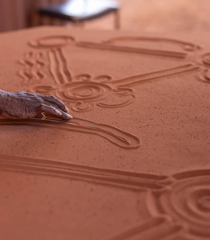 A close-up shot of an Aboriginal person's hand drawing traditional circular and linear symbols into a smooth layer of vibrant red sand.