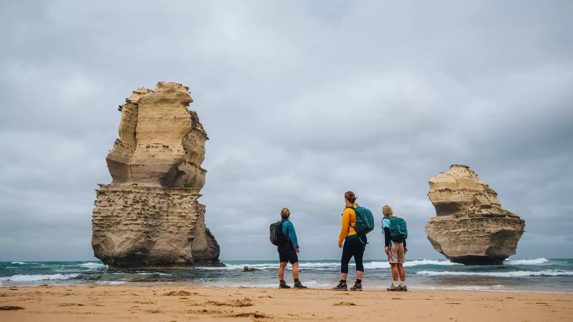 Three people standing on a sandy beach looking up at massive limestone stacks in the sea.
