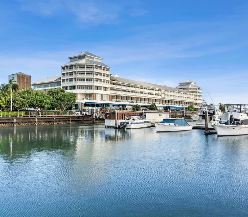 Interior view of a white hotel room with a king bed and glass doors opening to a balcony overlooking a marina in Cairns.