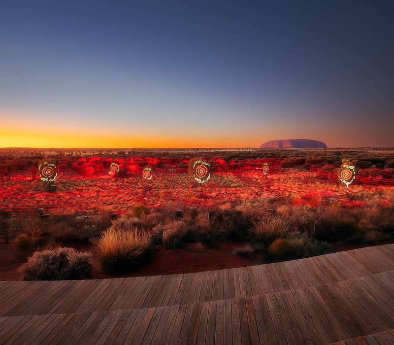 A wide, elevated view of a desert landscape at sunrise or sunset. The sky transitions from a deep blue at the top to a bright orange and yellow on the horizon. In the distance, the large silhouette of Uluru is visible.