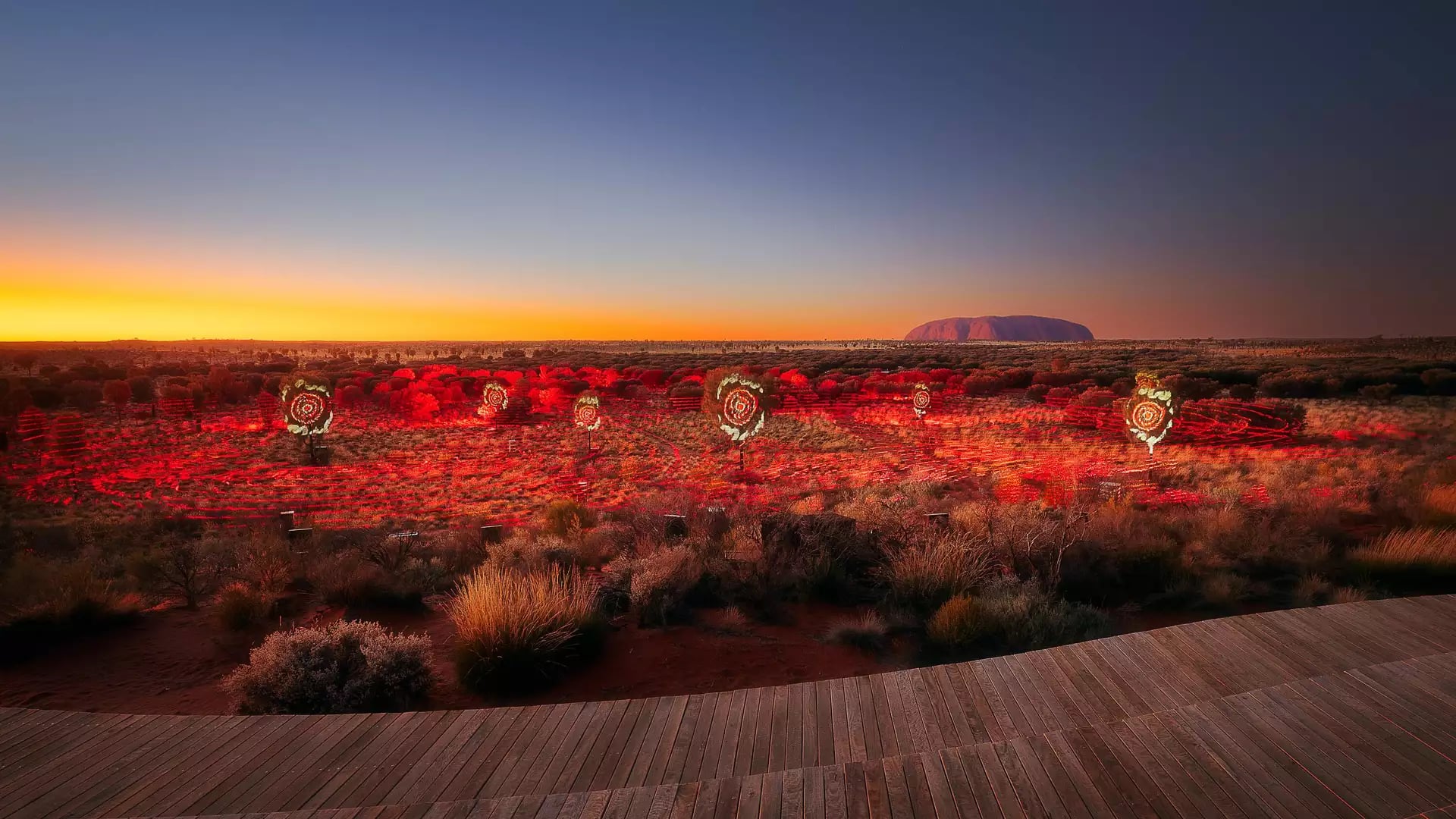 A wide, elevated view of a desert landscape at sunrise or sunset. The sky transitions from a deep blue at the top to a bright orange and yellow on the horizon. In the distance, the large silhouette of Uluru is visible.