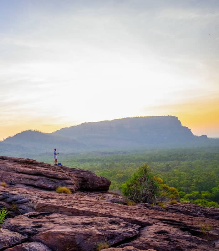 A photographer stands atop the rugged ironstone slopes of Nawurlandja Lookout, capturing the sunset glow over Anbangbang Billabong and the distant Nourlangie rock massif.