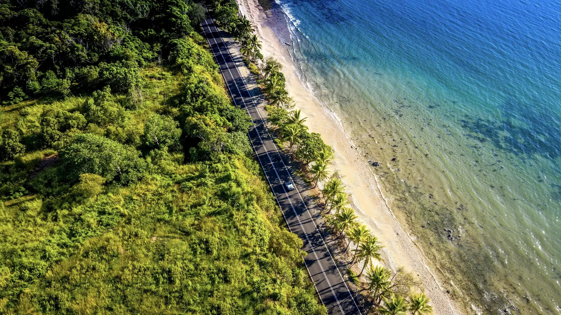 A high-altitude aerial view captures a white car traveling along a narrow asphalt road sandwiched between dense green tropical forest and a golden beach with turquoise water near Port Douglas.