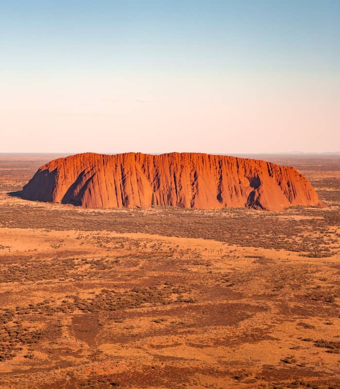 Aerial perspective of Uluru monolith surrounded by desert shrubland under a clear blue sky in the Red Centre.