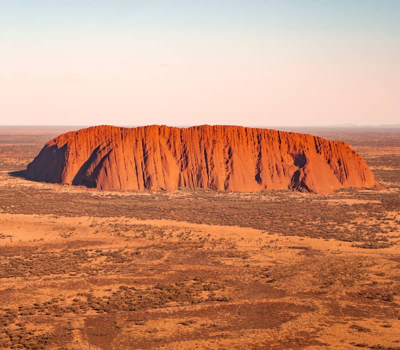 Aerial perspective of Uluru monolith surrounded by desert shrubland under a clear blue sky in the Red Centre.