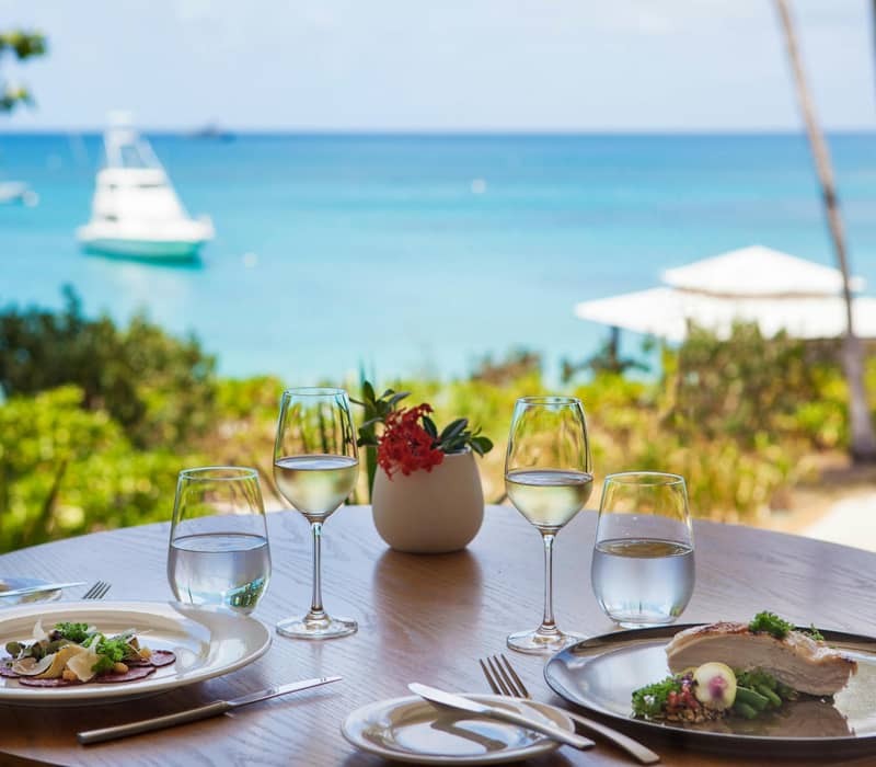Table with gourmet dishes and wine glasses by a window showing ocean and a yacht.