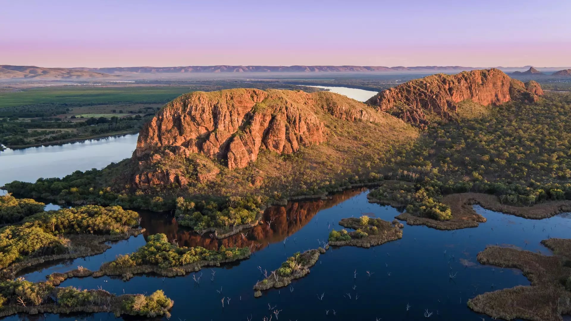 High-angle aerial view of a winding river and sunlit rocky ridges in the Kimberley at dawn.