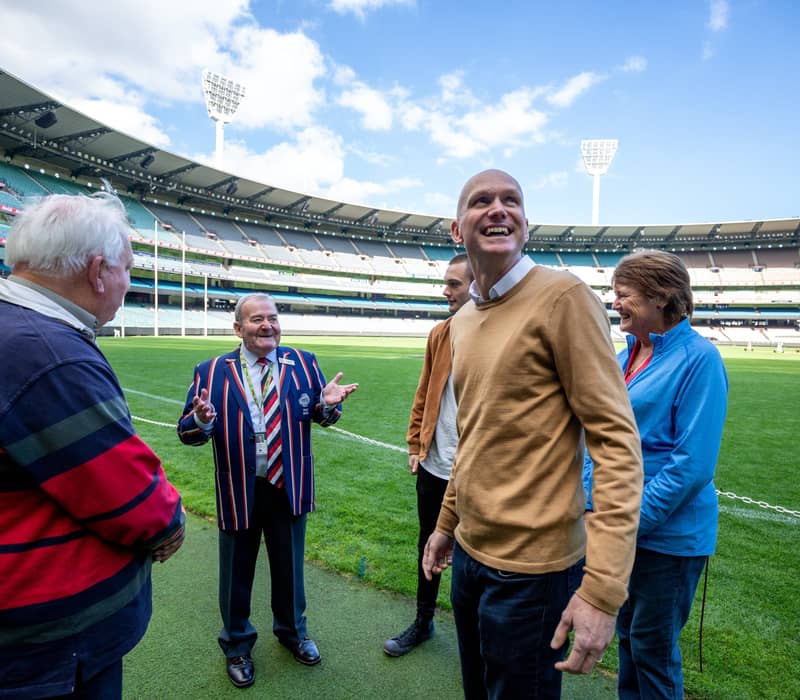 Visitors smiling and talking with a guide while standing on the grass at the Melbourne Cricket Ground.