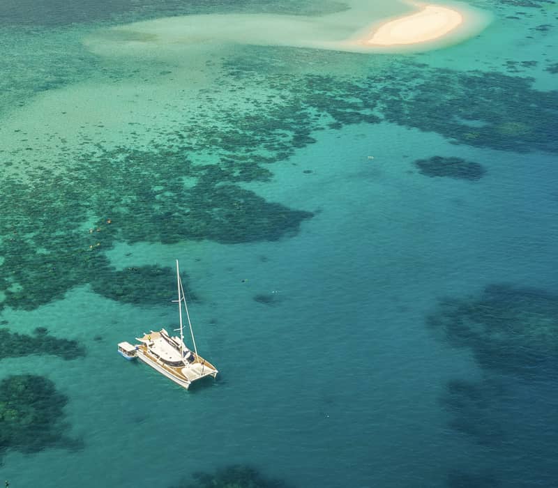 A high-angle drone shot captures a white sailing boat anchored in shimmering turquoise water next to a small, pristine white sandbar in the Great Barrier Reef.
