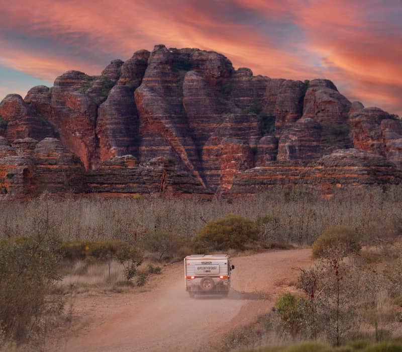 A rear view of a white caravan kicking up dust on a dirt track. In the background, large striped sandstone domes rise under a dramatic red and orange sunset sky.