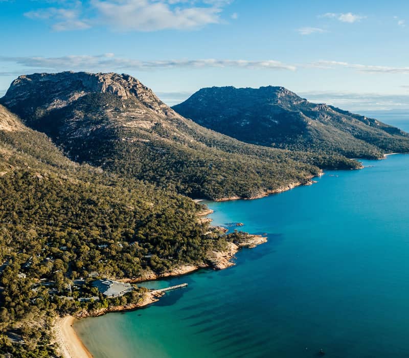 erial panorama of the Freycinet Peninsula coastline, featuring the main complex of Freycinet Lodge tucked into the dense green forest at the water's edge of Great Oyster Bay, with the striking pink granite peaks of the Hazards mountain range rising in the background.