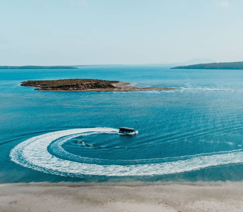A black boat makes a sharp circular turn in bright blue water, leaving a white wake near Coffin Bay