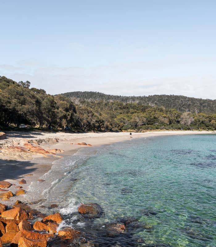 A view of Honeymoon Bay at Freycinet National Park, showing the clear turquoise water and a small sandy beach cove framed by orange granite boulders and native green trees under a soft, blue sky.