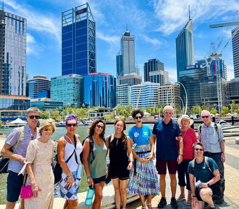 A group of tourists standing on the waterfront at Elizabeth Quay with skyscrapers and a blue sky behind them.