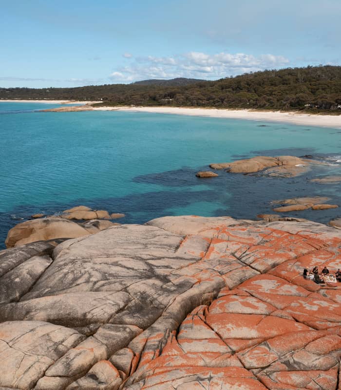 High-angle view of a group sitting on orange-stained rocks next to a white sand beach and turquoise ocean.