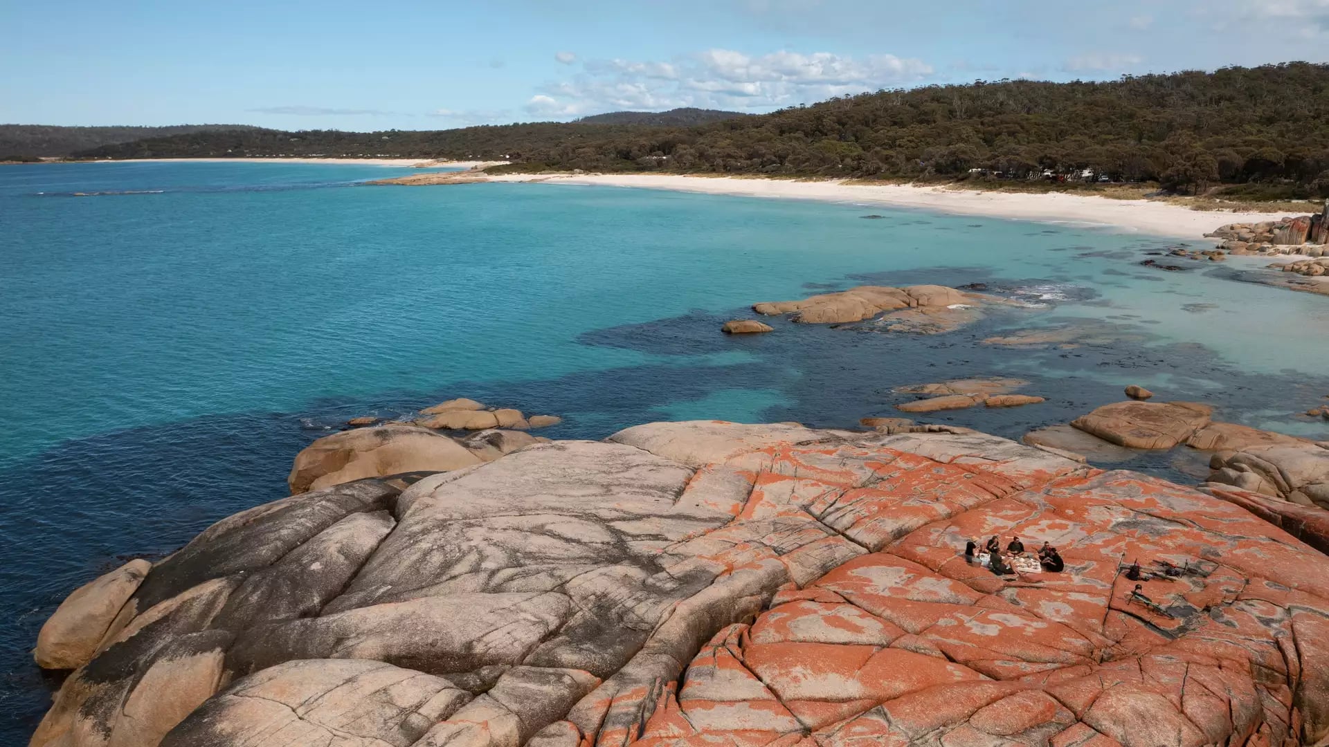 High-angle view of a group sitting on orange-stained rocks next to a white sand beach and turquoise ocean.