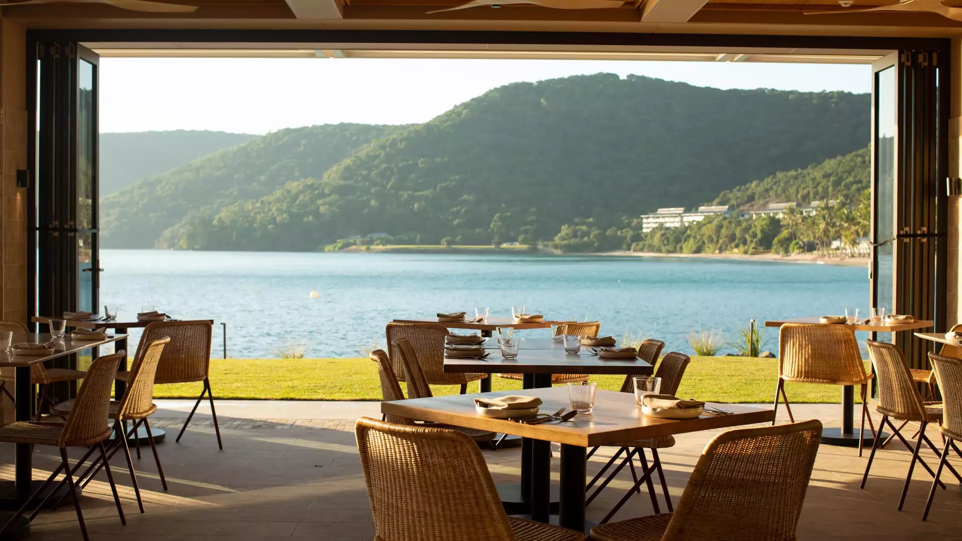 A restaurant interior with woven chairs and wooden tables looking out through open bi-fold doors to a calm blue bay and green hills.