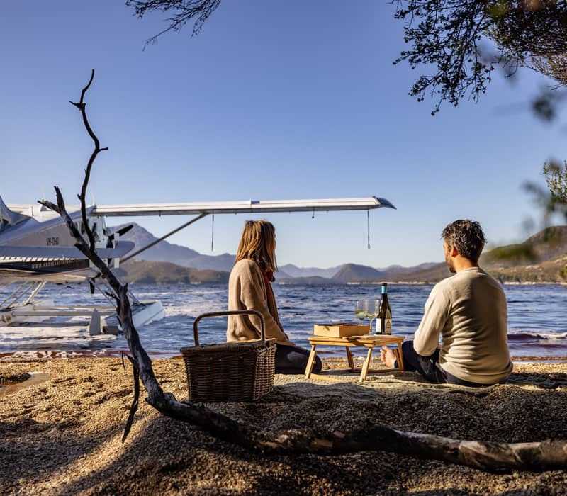 A couple enjoys wine and a picnic on a pebble beach with a seaplane in the background