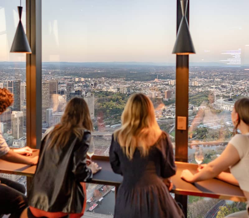 Four people standing at a floor-to-ceiling window overlooking the Melbourne city grid and sporting precinct.