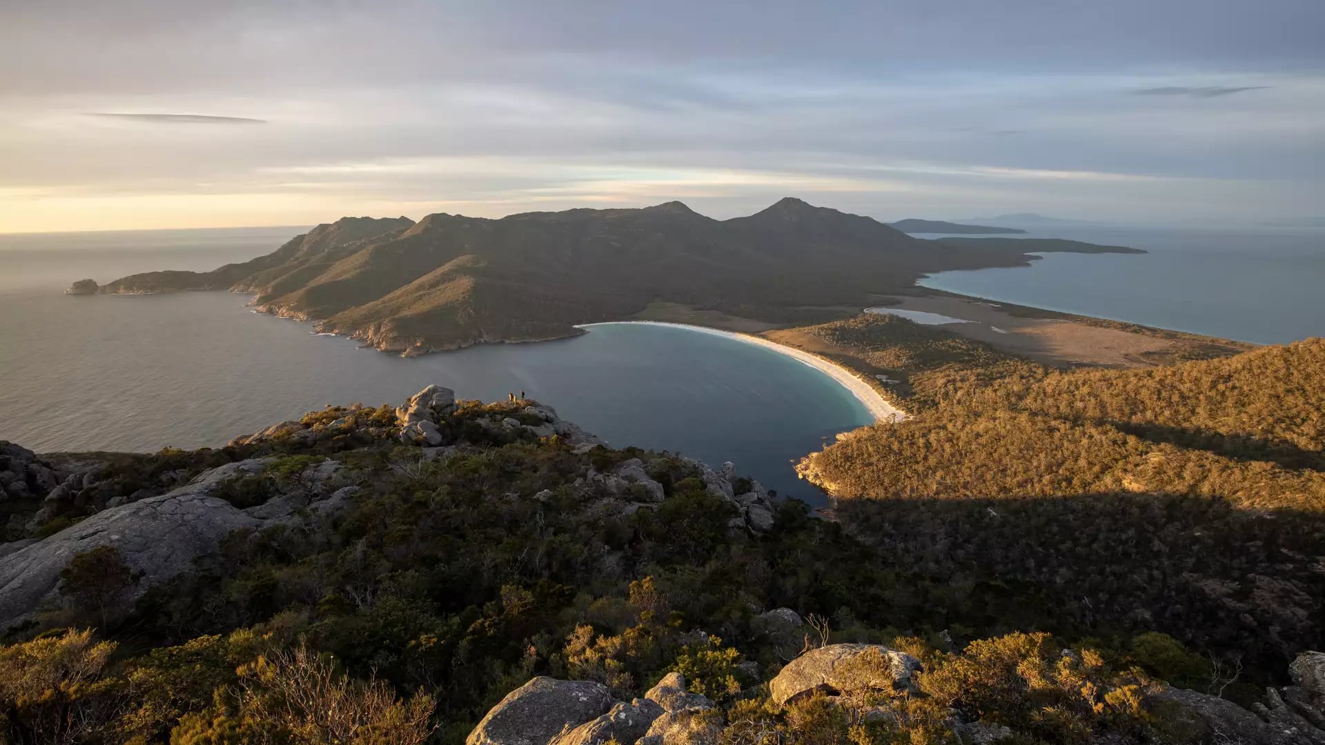 Panoramic high-angle view of a crescent-shaped white sand beach and turquoise bay surrounded by forested mountains under a soft sunrise sky.