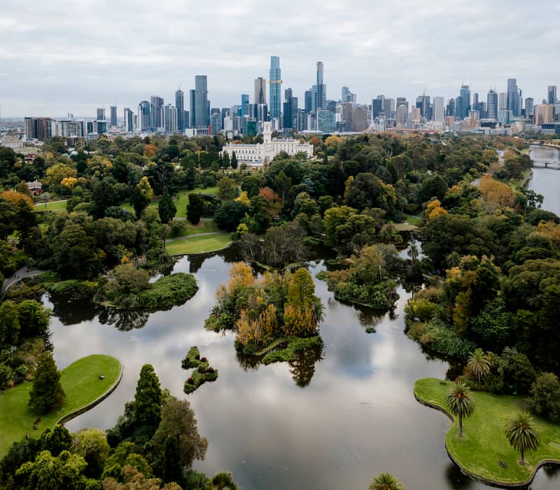 An aerial view of the Royal Botanic Gardens in Melbourne, Australia, showing a large lake, lush greenery, a white building, and the city skyline in the distance under a grey sky. 