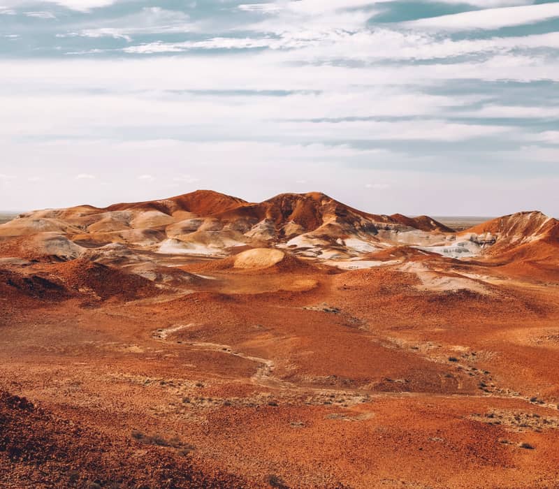 Vast desert landscape of the Coober Pedy Breakway's undulating hills of red, orange, and white earth under a pale blue sky with wispy clouds during The Ghan's journey