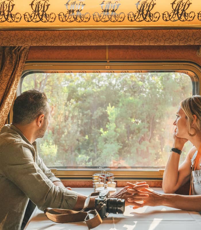 Interior view of a Gold Service Lounge on a Journey Beyond rail journey. A man and woman sitting at a white-clothed dining table inside a luxury Journey Beyond train carriage, looking out the window at green bushland.