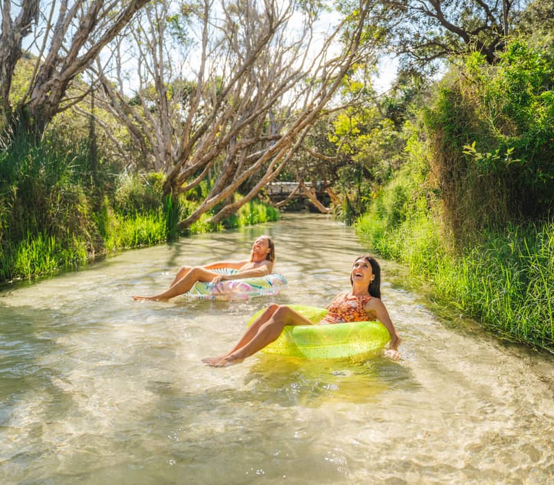 A man and woman on inflatable tubes floating down a clear forest stream on K'gari.