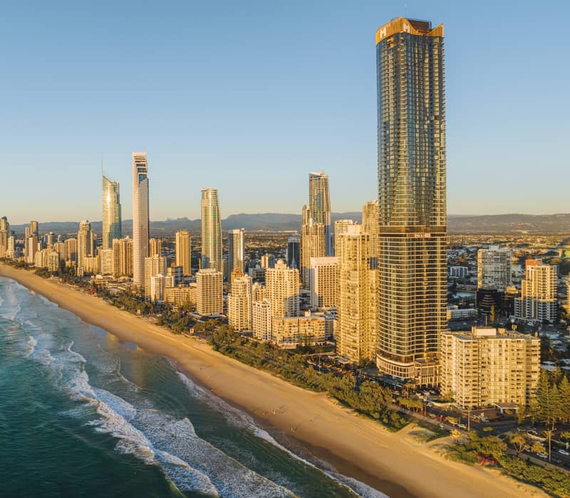 An aerial view of the Gold Coast skyline and Surfers Paradise beach with ocean waves during a golden sunset.