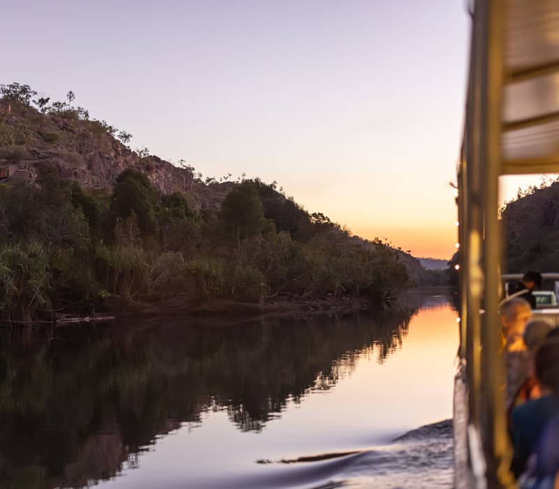 An aerial drone shot follows a white tour boat as it cruises down the center of a wide, calm river flanked by dense green bushland under a cloudy sky.