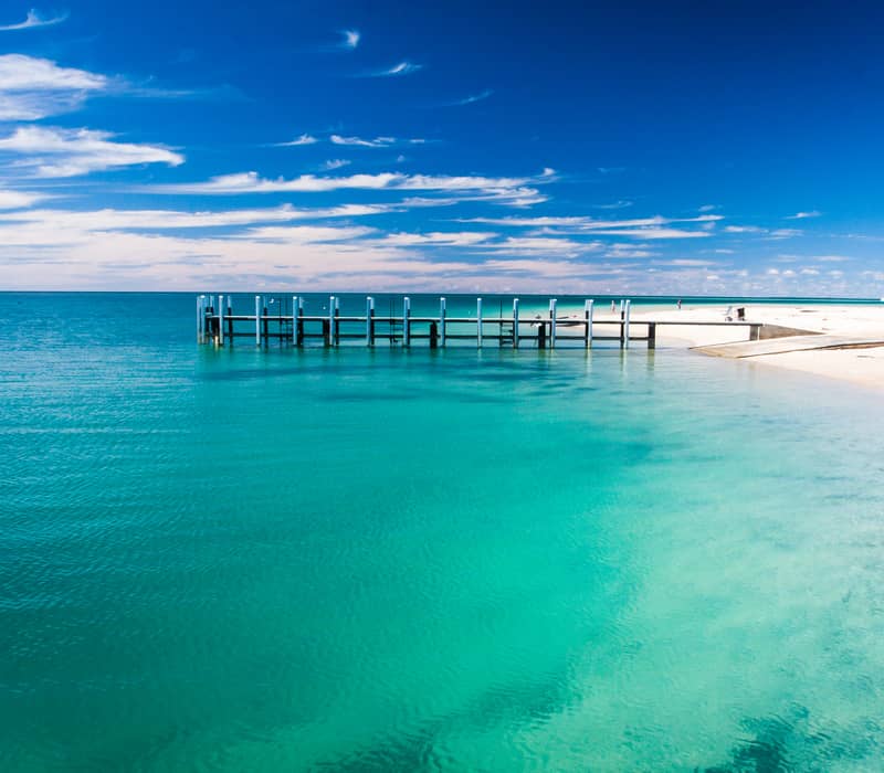 A white wooden jetty extends into vibrant turquoise ocean water from a white sand beach under a deep blue sky with wispy white clouds.