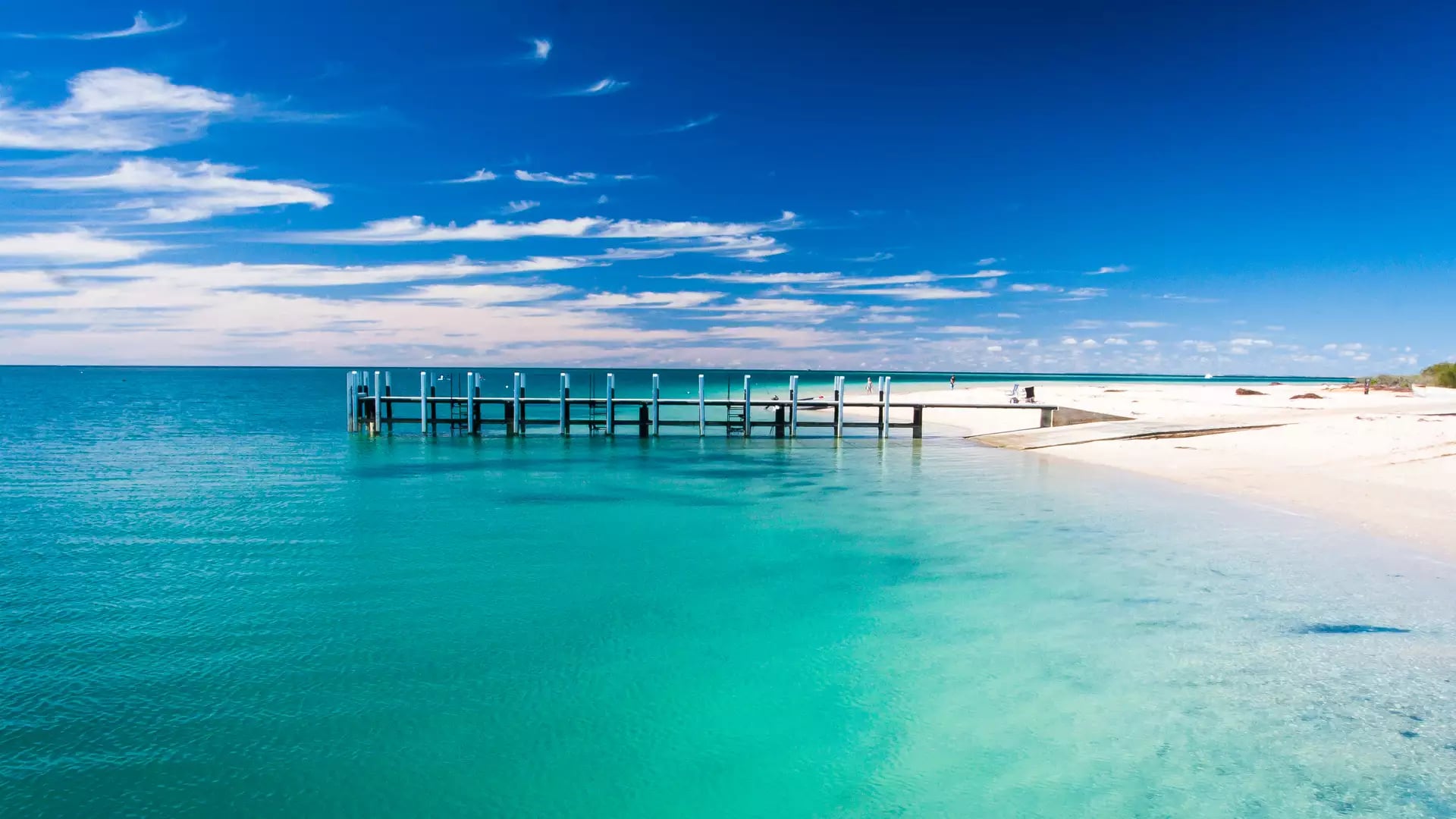 A white wooden jetty extends into vibrant turquoise ocean water from a white sand beach under a deep blue sky with wispy white clouds.
